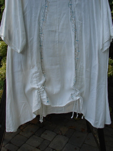 Barclay Linen Venetian Tunic Dress: A white shirt on a rack with blue embroidery. Feminine shape, A-line, double front draw cords, high side vents. Made from mid-weight linen. Size 1.