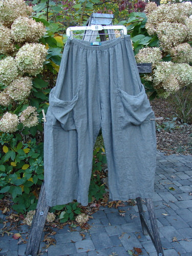 Barclay Lightweight Linen Side Pocket Pant Unpainted Army Stone Size 2, featuring an elastic waistband, wide legs, and large side pockets, displayed outdoors on a hanger against a patio and flowering shrubs.