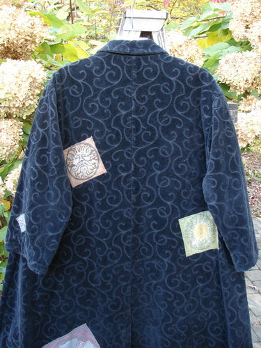 1998 Patched Tapestry Coat Bountiful Sun Guy Black OSFA: A rich, textured coat with curlicue patterns, three mandala-like patches, large pockets, and a stand-up collar, displayed among flowering shrubs.