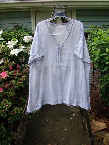 Barclay Batiste Deep Neck Tie Dress Unpainted Lilac Size 2 displayed on a clothes rack, showcasing its short sleeves and unique design detail of a slight light blemish on the lower single sleeve.