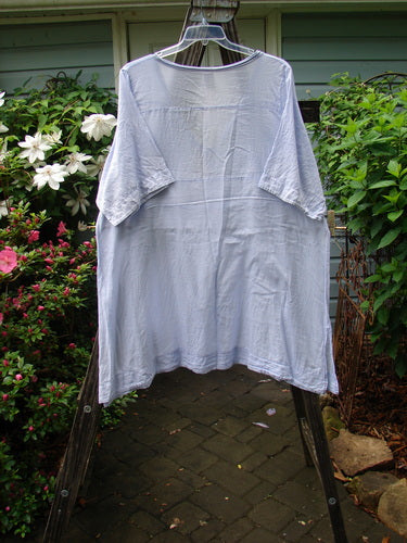 Barclay Batiste Deep Neck Tie Dress Unpainted Lilac Size 2 displayed on a clothes hanger on a rack, showcasing its short sleeves and slight light blemish on a single lower sleeve.