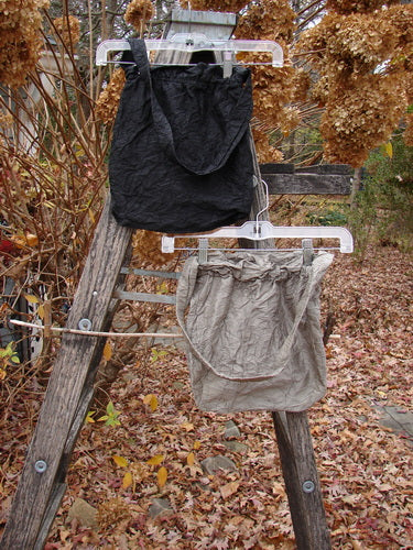 Two 2000 NWT Shaunting Silk Bags in Unpainted Black and Cement hang from a ladder, showcasing their crinkled silk texture, medium shoulder straps, and elastic-topped closures amid an autumnal outdoor setting.