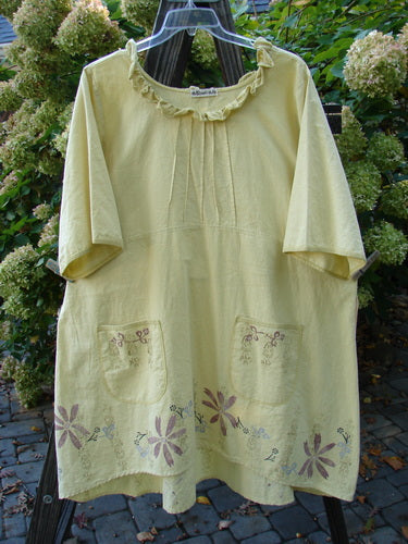A Barclay Linen Duet Sunrise Dress in Sunshine, size 2. A sweet baby doll tunic with a curvy neckline, pleated bodice, and a widening shape. Features include a slight empire waistline, front pockets, and a tiny floral and Roman goddess theme. Made from a unique cotton linen fabric.