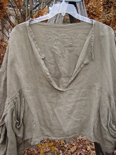 Magnolia Pearl Linen Crop Artist's Pocket Pullover Light Army OSFA displayed outdoors, showcasing its deep V-neck, three-quarter billowy sleeves, cropped wide shape, varying hemline, and dual oval pockets.