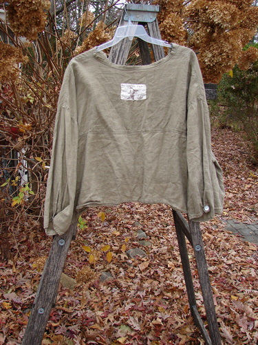 The Magnolia Pearl Linen Crop Artist's Pocket Pullover Light Army OSFA is displayed on a wooden ladder, showcasing its billowy three-quarter sleeves and unique pocket design against an autumn backdrop.