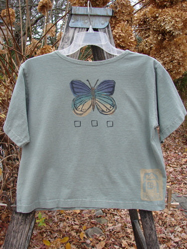 1996 Crop Tee Floral Butterfly Cricket Size 1 displayed on a hanger over a wooden ladder, features a blue and green butterfly graphic with check-boxes, showcasing a vintage style from Bluefishfinder.com.