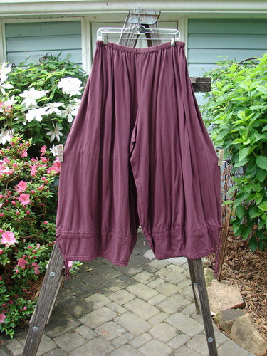 A pair of Barclay Tie Bottom Square Pants in Unpainted Maroon with a replaced elastic waistline and oversized pockets, displayed on a wooden rack.