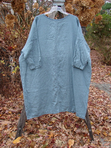 Barclay Linen Oversized Gallery Dress Falling Daisy Delaware OSFA hangs on a wooden hanger, showcasing its loose fit with a rounded neckline, drop shoulders, and three-quarter dolman sleeves amidst an autumn garden.