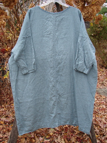 Barclay Linen Oversized Gallery Dress Falling Daisy Delaware OSFA: A slate-blue tunic-style dress with a rounded neckline, dropped shoulders, and elbow-length dolman sleeves, hanging outdoors on a wooden hanger.