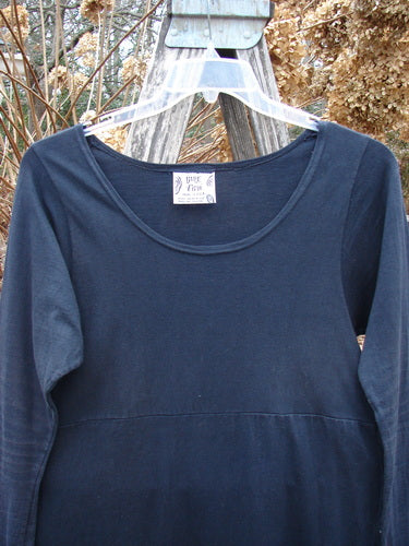 1992 Pedestal Dress Unpainted Black Size 1, hanging on a weathered wooden support, showcases a deeper rounded neckline and empire waist, with unique fishtail side flutters, reflecting vintage elegance.