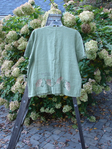 1998 Botanicals Robin's Top, a green shirt on a wooden rack. Features include a smaller back shoulder line, elastic-topped pockets, and perfect buttons. Made from heavy-weight linen. Bust 46, Waist 48, Hips 50, Hem Circumference 54, Length 27.