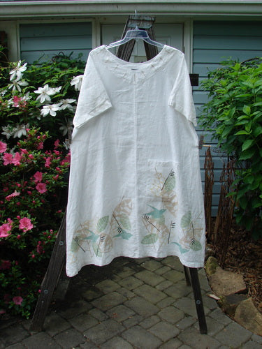 A white Barclay Linen Vertical Seam Pocket Dress with leaf and dove theme, displayed on a hanger outdoors. The dress features a rounded neckline, wide short sleeves, and a front pocket.