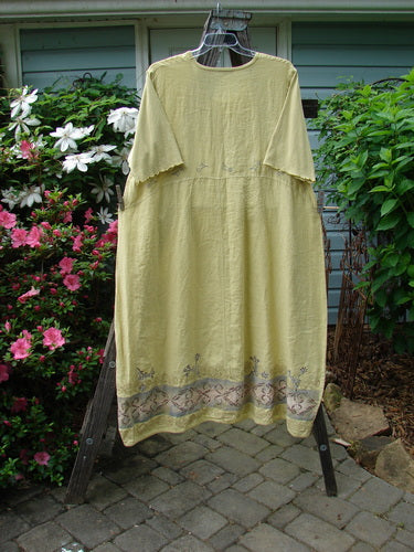A Barclay Linen Cotton Sleeve Banded Hem Pleat Dress in Plantain displayed on a rack, featuring vertical pleats, a downward curved empire waist, and three-quarter length sleeves.