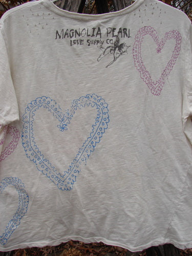 Magnolia Pearl NWT Cotton Jersey Boyfriend Tee Frida Del Sol True OSFA displayed outdoors, featuring lace-style hearts and MAGNOLIA PEARL LOVE SUPPLY CO. text near the neckline, showcasing unique design elements and distressing.