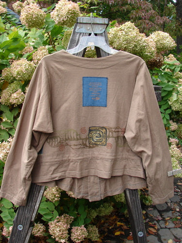 Image: A brown shirt with a blue square on it, featuring a close-up of a bush, a blue rectangular object with text, a close-up of a wood plank, a close-up of a fish tail, and a close-up of a flower.

Alt text: 1997 Treehouse Jacket Primitive Climb Lumber Size 1: A brown shirt with a blue square on it, surrounded by nature elements like a bush, a wood plank, a fish tail, and a flower.
