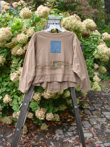 Image: A brown shirt on a wooden stand, with a close-up of a metal object.

Alt text: 1997 Treehouse Jacket in Lumber, Size 1, on wooden stand. V-neck, button line, drawcord flounce. Blue Fish patch. Bust 50, waist 58, front length 26, sleeves 28, back length 21.