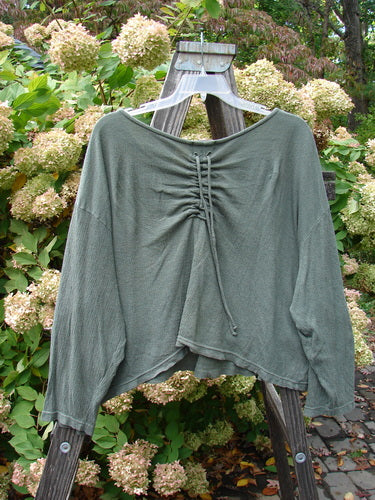2000 Limited Edition Gauze Ruched Back Top Unpainted Green Lake Size 2: A long-sleeved green shirt on a wooden rack, with a close-up of a plant and a screw on a wood post.