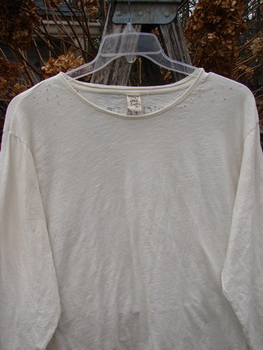 Magnolia Pearl Cotton Jersey Long Sleeved Boyfriend True OSFA shirt on hanger, displaying subtle eyelet details at shoulders and neckline, with drapey feel and upper shoulder distressing, set against wooden backdrop.