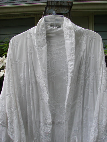 A white Barclay Brocade Open Front Pocket Dress Duster with floral vine patterning, displayed hanging outdoors. Features include drop exterior pockets, varying hemline, and fold-over collar. Size 2.