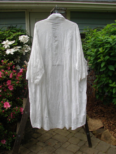 Alt text: Barclay Brocade Open Front Pocket Dress Duster in white, displayed on a hanger, featuring exterior pockets, floral vine pattern, and varying hemline.