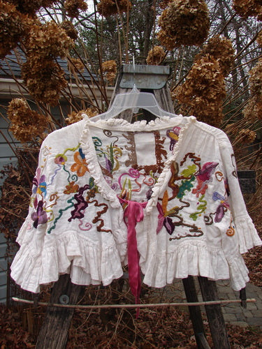 Magnolia Pearl Embroidered Linen Ruffle Ribbon Bolero White OSFA features intricate floral embroidery with ruffled edges and a pink satin ribbon tie, draped elegantly over a rustic wooden ladder.