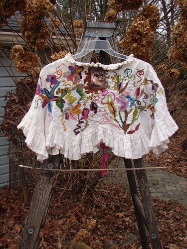 Magnolia Pearl Embroidered Linen Ruffle Ribbon Bolero White OSFA draped over a wooden ladder, showcasing intricate multicolored floral embroidery, scalloped ruffles, and a unique sewn image, reflecting artistic craftsmanship.