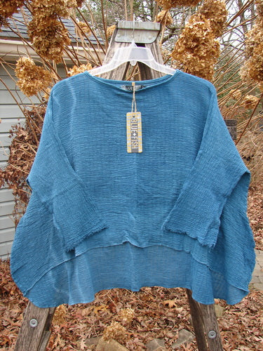 The Barclay NWT Crinkle Gauze Top in Mottled Teal OSFA, displayed on a hanger, features a loose-fitting, textured design with a high-low hemline and three-quarter dolman sleeves.