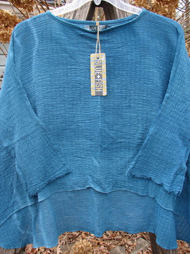 Barclay NWT Crinkle Gauze Top Mottled Teal OSFA, featuring a crinkled texture, high-low hemline, and dolman sleeves, displayed on a hanger with a Blue Fish tag, against dried leaves backdrop.