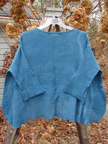 Barclay NWT Crinkle Gauze Top in Mottled Teal OSFA, featuring a boxy silhouette with wide three-quarter dolman sleeves, frayed edges, and a high-low hemline, displayed on a hanger outdoors.