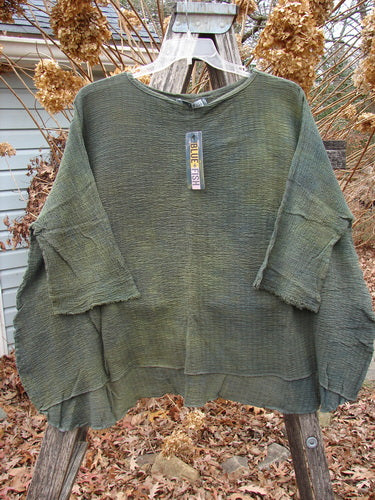 Oversized Barclay NWT Crinkle Gauze Top in Mottled Evergreen displayed outdoors on a wooden trellis, showcasing its loose-knit texture, high-low hemline, and three-quarter dolman sleeves amidst autumnal hydrangeas.