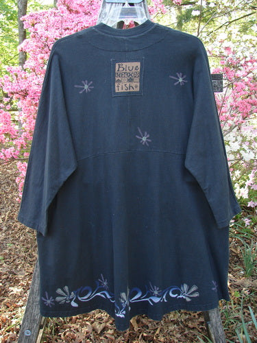1997 Rhadha Dress Star Flowers Trail Obsidian Size 2: A vintage black tunic with embroidered starburst motifs and floral hem, featuring a mock Mandarin collar and Blue Fish label, displayed against a garden backdrop.