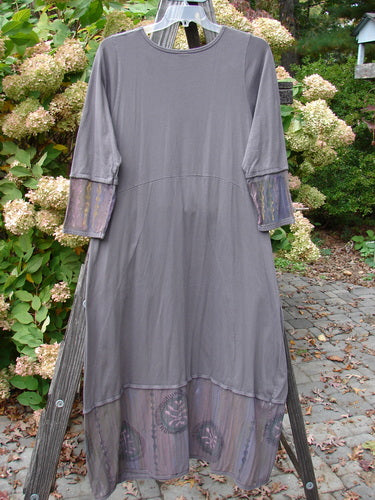 A long grey dress with a unique pleated front neckline, sectional painted lover sleeves, and a sweeping A-line shape. It features double exterior mommy drop pockets, a generous banded bottom sectional panel, and a downward curved empire waist seam. Painted in detailed fern graphics. Size 1.
