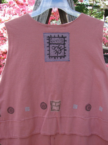 1995 Button Vest Village Russet OSFA displayed outdoors, featuring a dusty-rose color, square blufish patch, hand-drawn shapes, and unique hemline against pink flowering trees, embodying vintage Blue Fish style.