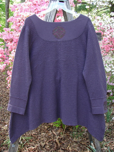 Barclay Patched Crepe Cross Over Tunic Top Diamond Brum Size 1, featuring a textured knit with a unique cross-over neckline, asymmetric hem, and diamond-themed patches, displayed on a wooden ladder amidst flowering shrubs.