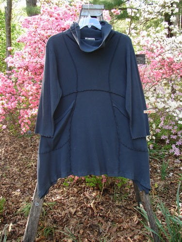 Barclay Thermal Reverse Stitch Pocket Dress Unpainted Raven Size 1 on wooden rack, highlighting its cowl neck, angled hemline, and front pockets, with a backdrop of flowering shrubs.