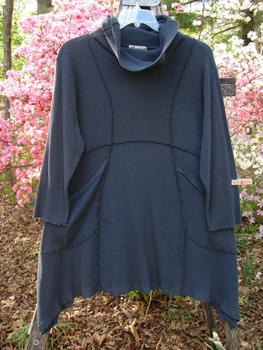 Barclay Thermal Reverse Stitch Pocket Dress Unpainted Raven Size 1, featuring a wide draped collar, exposed seams, and oversized pockets, displayed on a hanger amidst flowering shrubs.