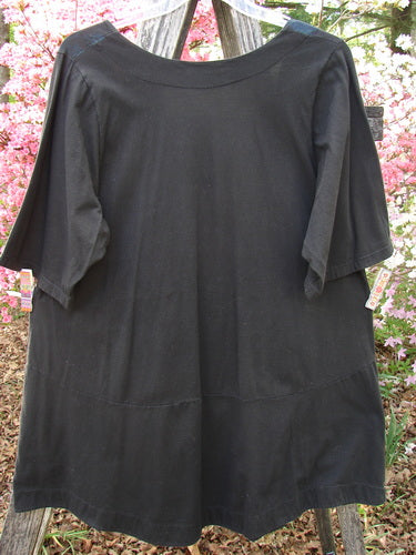 The Barclay Short Sleeved Be There Top Floral Bud Black Size 1 elegantly drapes over a wooden stand, showcasing its flowing silhouette and unique neckline. It features angular pockets and a gracefully banded hemline.