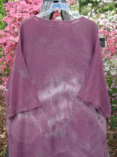 Barclay Crinkle Gauze Ana Tunic Dress Unpainted Mottled Rusty Rose OSFA displayed on a hanger, showcasing its loose fit, wide three-quarter sleeves, and varying hemline, with floral shrubs in the background.