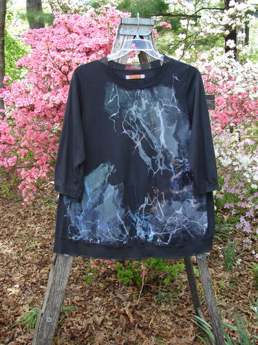Barclay Flutter Edged Essential Tee in Lightning Black, Size 1, features abstract pale blue splatters on three-quarter sleeves, mesh neckline, and hem. Displayed on a wooden ladder amidst blooming azaleas.