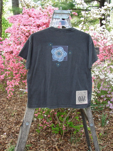 1997 Short Sleeved Tee Butterfly Ebony Size 0 draped on a wooden ladder, featuring a floral graphic on the back, Blue Fish Signature Patch, and a thick ribbed neckline, amidst blooming shrubs.