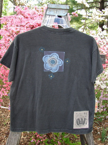 1997 Short Sleeved Tee Butterfly Ebony Size 0, draped over a wooden frame, features a large stylized flower motif on the back. Signature patch with text near the hem. Photographed outdoors.