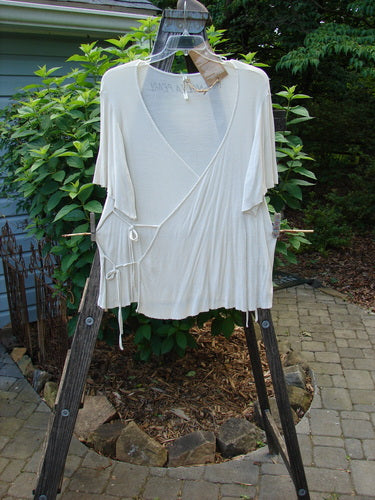 Alt text: Magnolia Pearl NWT Ribbed Origami Wrap Blouse Moonlight OSFA displayed on a wooden clothes rack outdoors. Features deep neckline, wide sleeves, varying hemline, and tie closures.