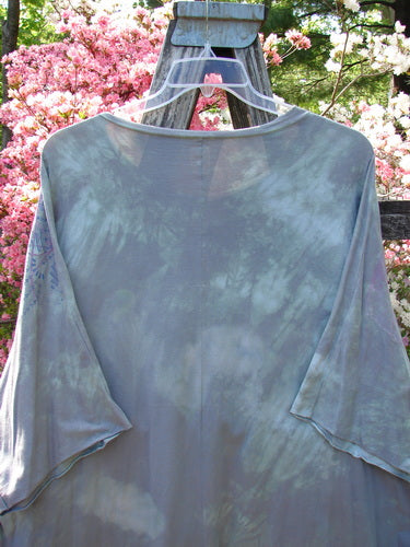 Barclay Flutter Collar Essential Tee Mood Garden Mottled Gray Grass OSFA on a hanger, featuring a flutter neckline, raw edge dolman sleeves, and hi-low hemline, displayed outdoors against floral shrubs.