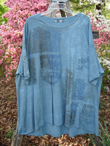 Barclay Art Tunic Tee Cosmic Celtic Steel Blue OSFA displayed outdoors, showcasing its loose fit, three-quarter sleeves, rounded neckline, and distinctive upward rounded front hem against a backdrop of flowering shrubs.