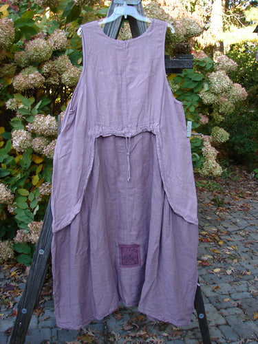 Barclay PMU Linen Explorer Jumper Pansy Lavender Size 2: A lavender dress on a wooden stand, featuring a scoop neckline, full bodice overlay, billowy lower sweep, and pansy theme paint.