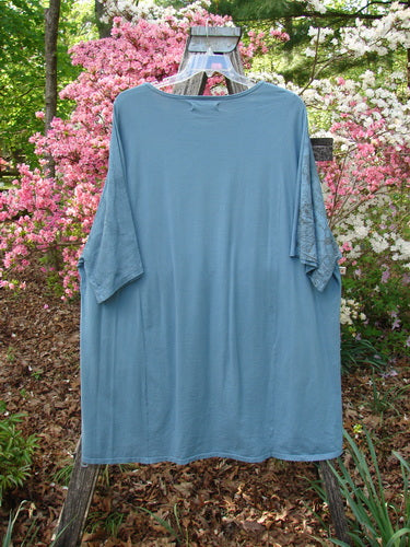 Barclay Art Tunic Tee Cosmic Celtic Steel Blue OSFA displayed on a wooden hanger, featuring a rounded neckline, drop shoulders, and loose three-quarter sleeves against a garden backdrop with blooming azaleas.