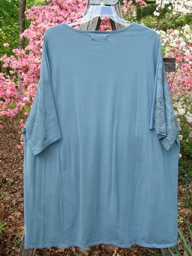 Barclay Art Tunic Tee Cosmic Celtic Steel Blue OSFA, featuring elbow-length sleeves with lace insets, hangs outdoors amidst blooming azaleas. Tunic showcases a rounded neckline and varying hemline.