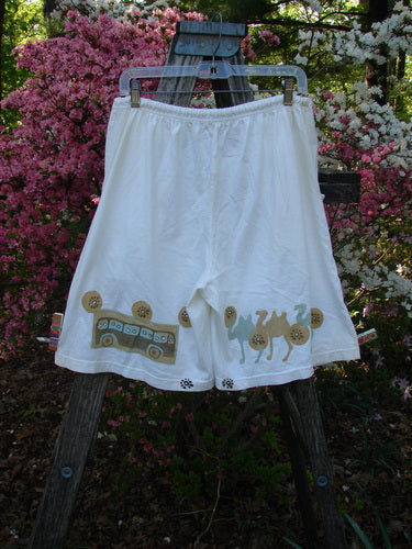 2000 Bushel Short Bus Travel Milk Size 1, featuring loose-fitting white cotton with travel-themed appliqué motifs, displayed on a hanger over a ladder, showcasing deep front pockets and a drawstring waist.