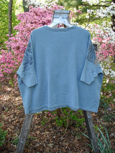 Barclay Fleece Banded Sleeve Dolman Top Moon Garden Sage OSFA draped on a wooden ladder, showcasing its wide boxy shape, dolman sleeves, and banded hem in a blooming garden setting.