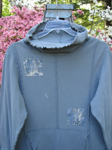 Barclay Patched Thermal Cowl Neck Roo Tunic Gray Day Size 1, showcasing decorative patches and exposed seams, hangs on a hanger outdoors, highlighting its cowl neck and ribbed waffle cuffs in a garden setting.